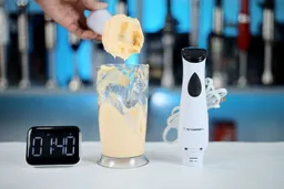 A hand pours a creamy mixture from a cup beside the Elite Gourmet immersion blender; a digital timer reads 01:40, set against a backdrop of kitchen appliances.