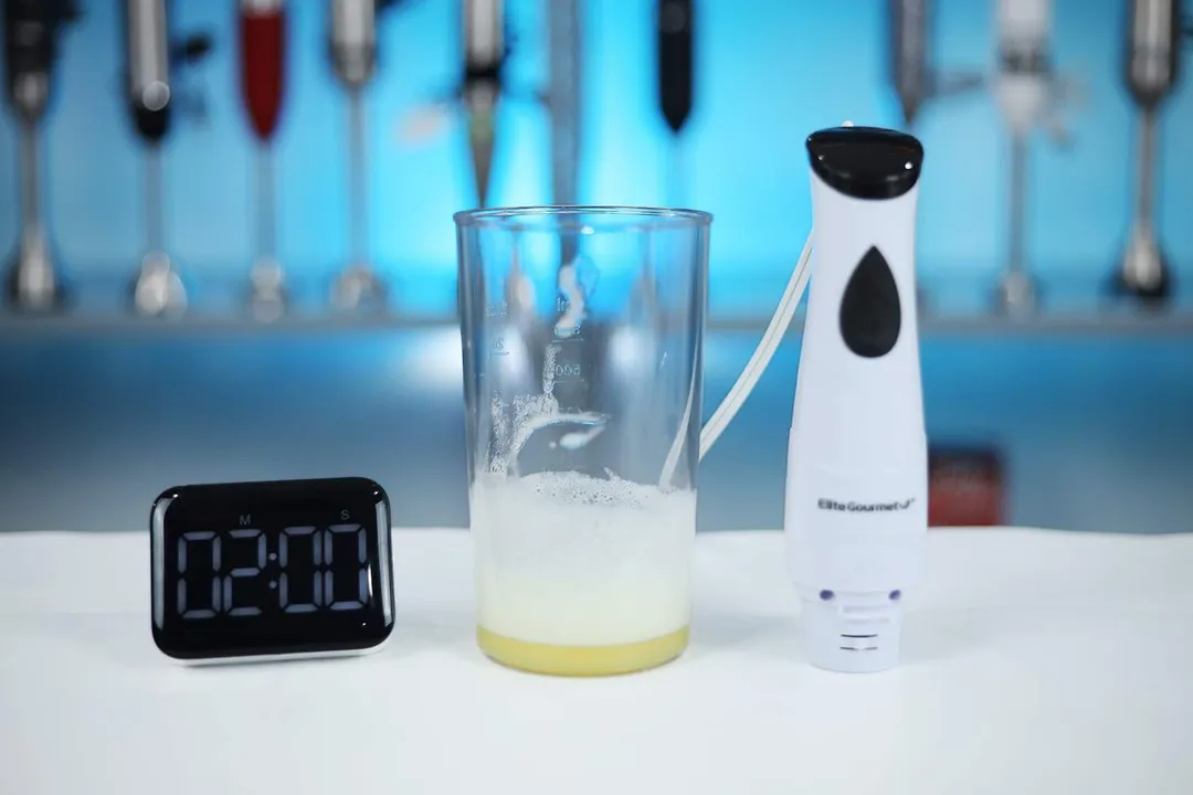 A digital timer shows 2:00 on a kitchen counter, with a beaker containing unsuccessfully beaten egg whites next to the Elite Gourmet immersion blender.
