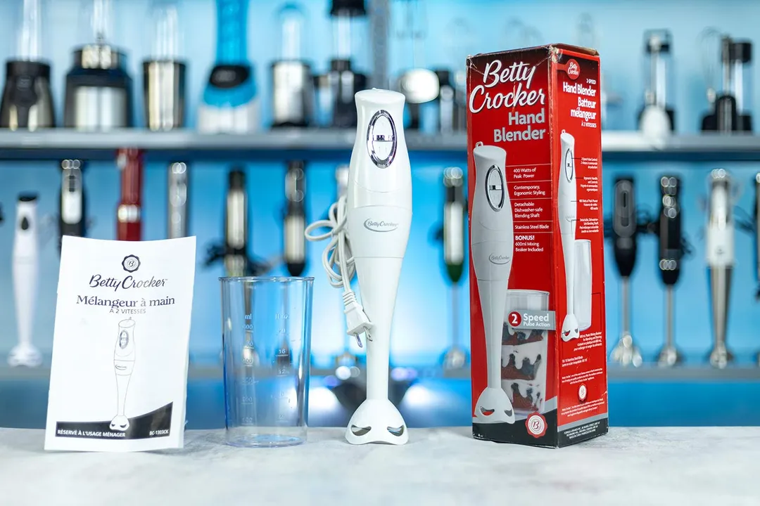The Betty Crocker BC-1303CK Immersion Blender standing on a table with a beaker, user manual, and packaging box by its sides.