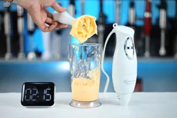 A hand pours a creamy mixture from a cup beside theBetty Crocker immersion blender; a digital timer reads 2:15, set against a backdrop of kitchen appliances.
