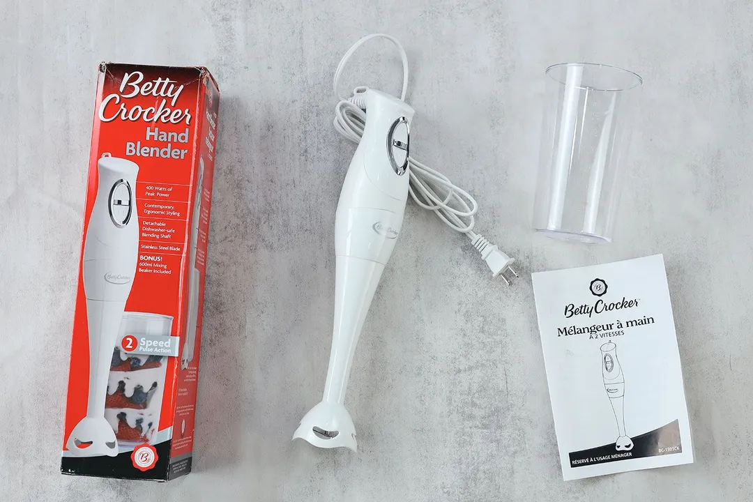 The Betty Crocker immersion blender lying on a table with its accessories include a beaker, user manual, and packaging box.