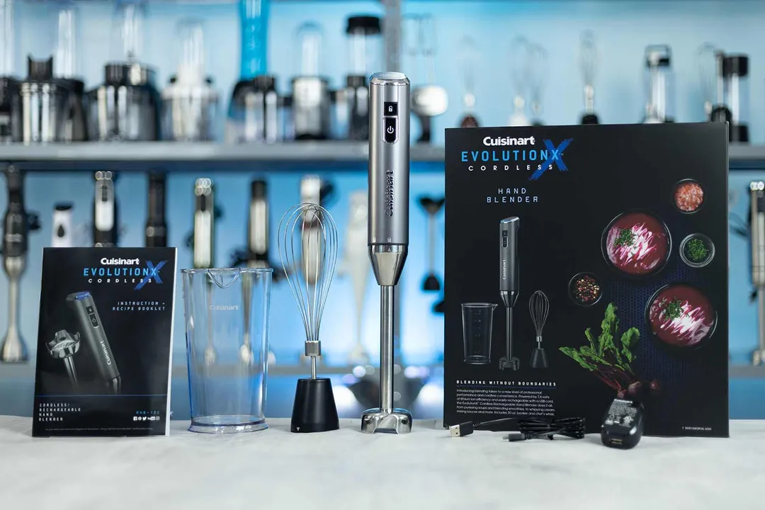 A Cuisinart EvolutionX Cordless hand Blender set on a kitchen counter, with a whisk attachment, a charging base, a recipe booklet, and the product packaging by its sides.