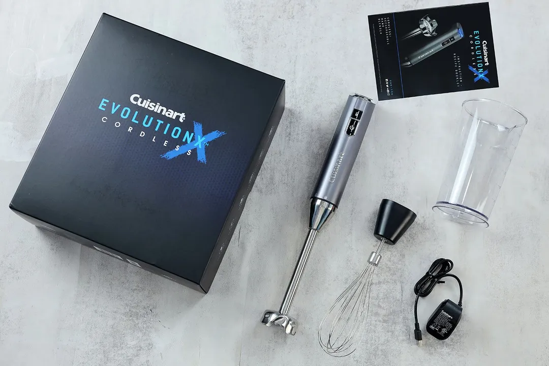 The Cuisinart EvolutionX RHB-100TG immersion blender lying on a table with its beaker, whisk attachment, user manual, packaging carton box, and charger by its side.