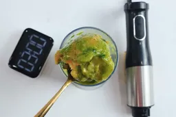 A hand holding a spoonful of a chunky, unevenly blended green and orange mixture above the LINKchef immersion blender, demonstrating the results of a failed frozen fruit smoothie test with the LINKchef blender.