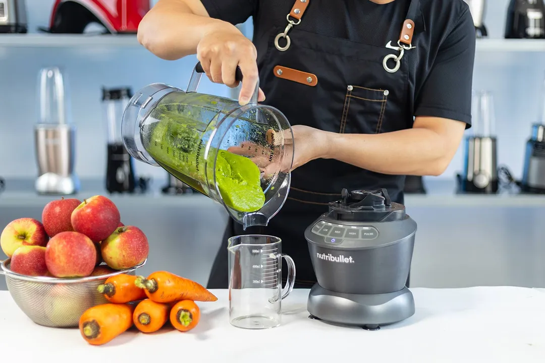 Someone pouring a green smoothie from a NutriBullet Blender Combo 1200 Watt into a glass, with apples and carrots on the countertop, and a selection of blenders on shelves behind.