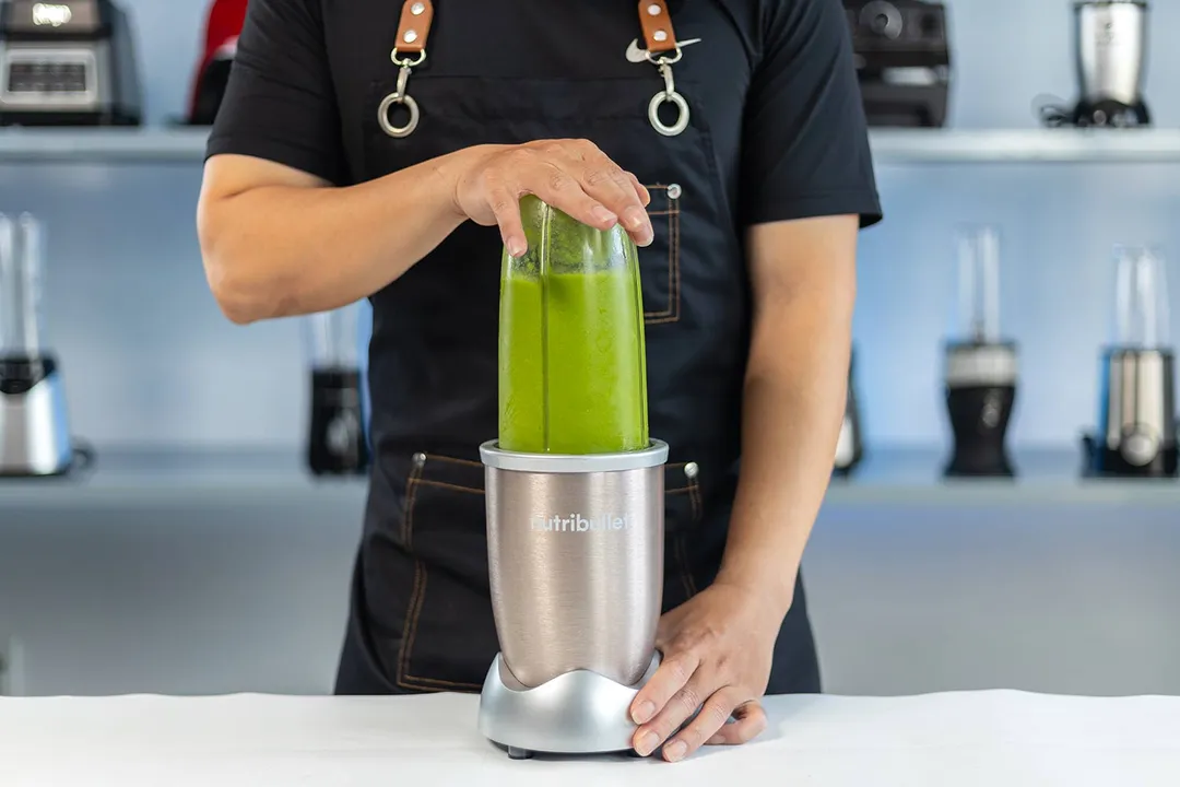 Someone securing a green smoothie-filled NutriBullet blender cup on the blender base, with kitchen appliances blurred in the background.