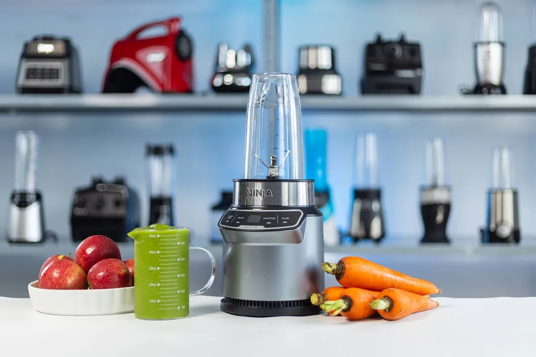 The Ninja Nutri Pro Compact Personal Blender (BN401) with a cup containing a green smoothie next to a bowl of apples and a bunch of carrots, with various blenders in the background.