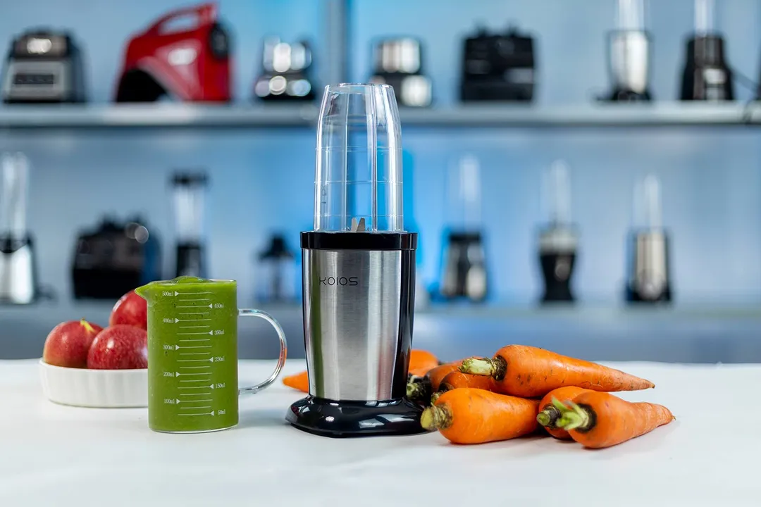 The KOIOS Pro Bullet personal blender with a cup containing a green smoothie next to a bowl of apples and a bunch of carrots, with various blenders in the background.