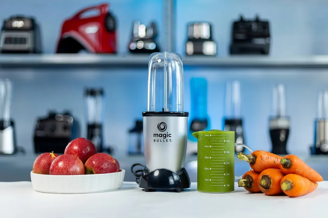 The Magic Bullet 250W personal blender with a cup containing a green smoothie next to a bowl of apples and a bunch of carrots, with various blenders in the background.