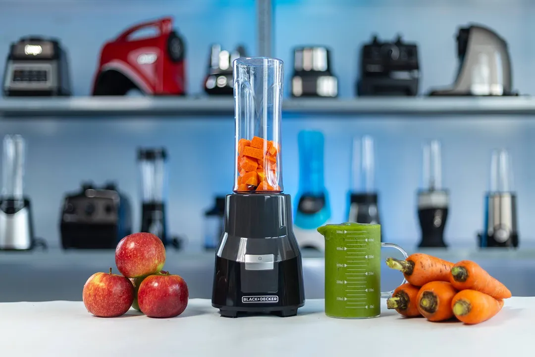 The Black+Decker personal blender with a cup containing a green smoothie next to a bowl of apples and a bunch of carrots, with various blenders in the background.