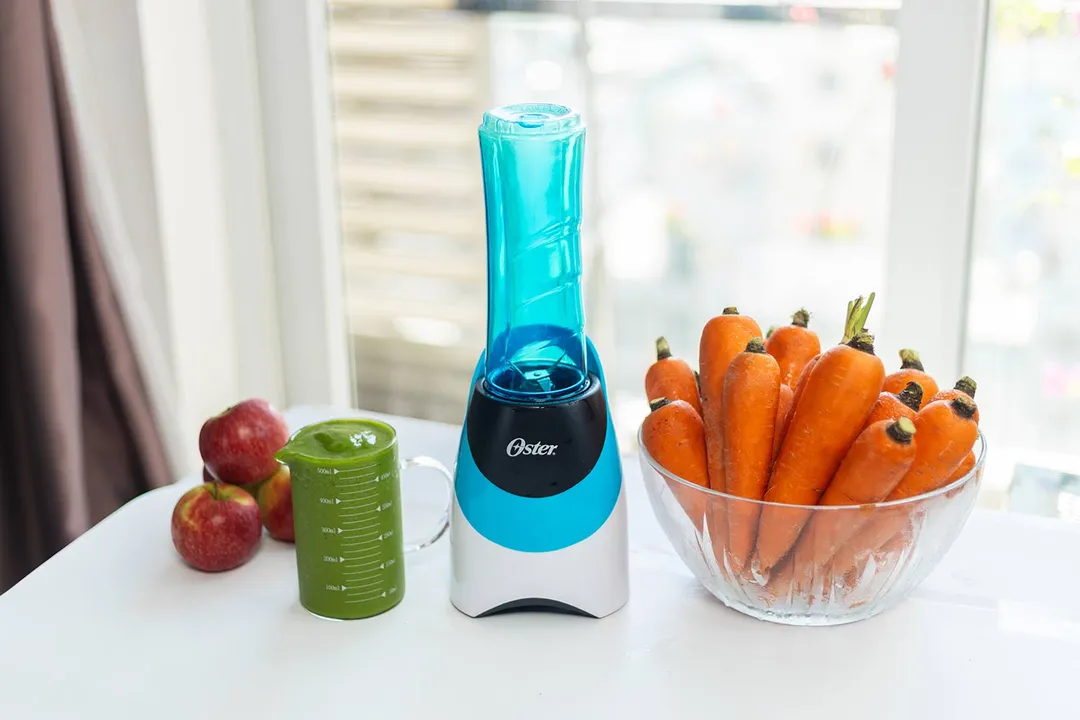 The Oster Pro personal blender with a cup containing a green smoothie next to a bowl of apples and a bunch of carrots.