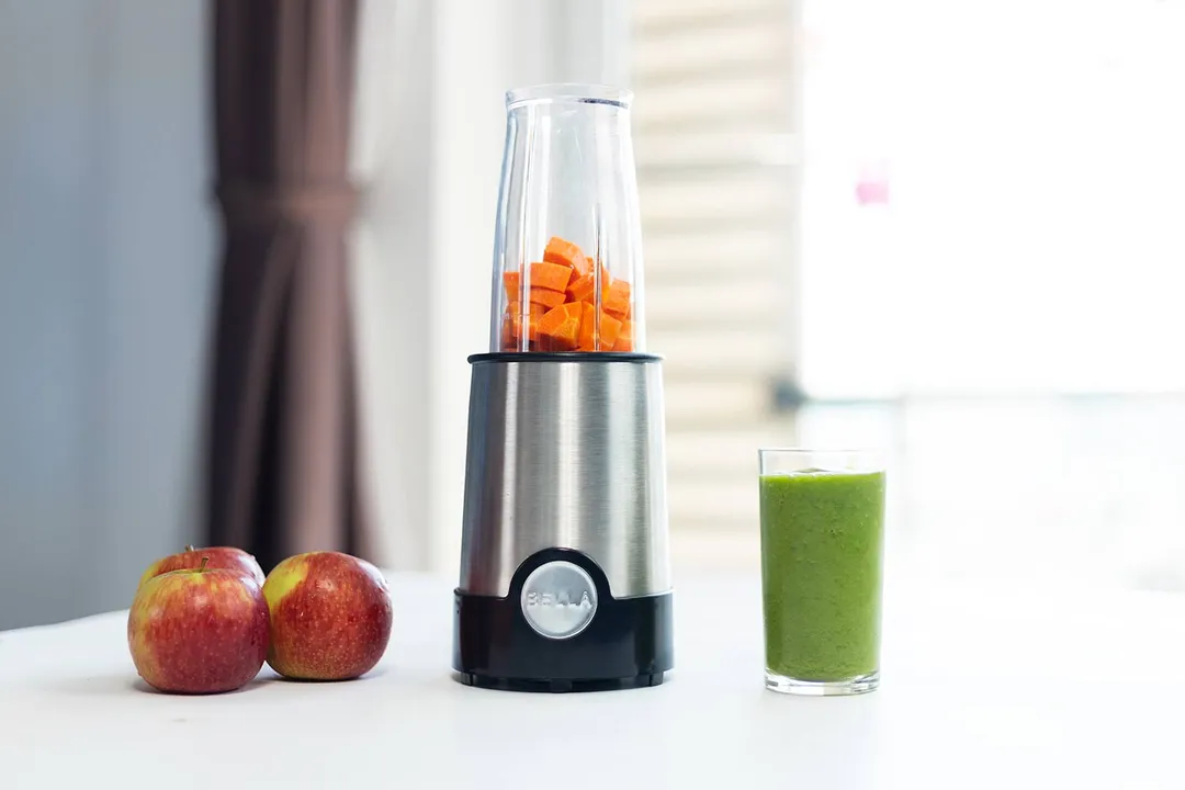The BELLA personal blender with chopped carrots in its cup and a green smoothie in a glass, alongside two whole apples.