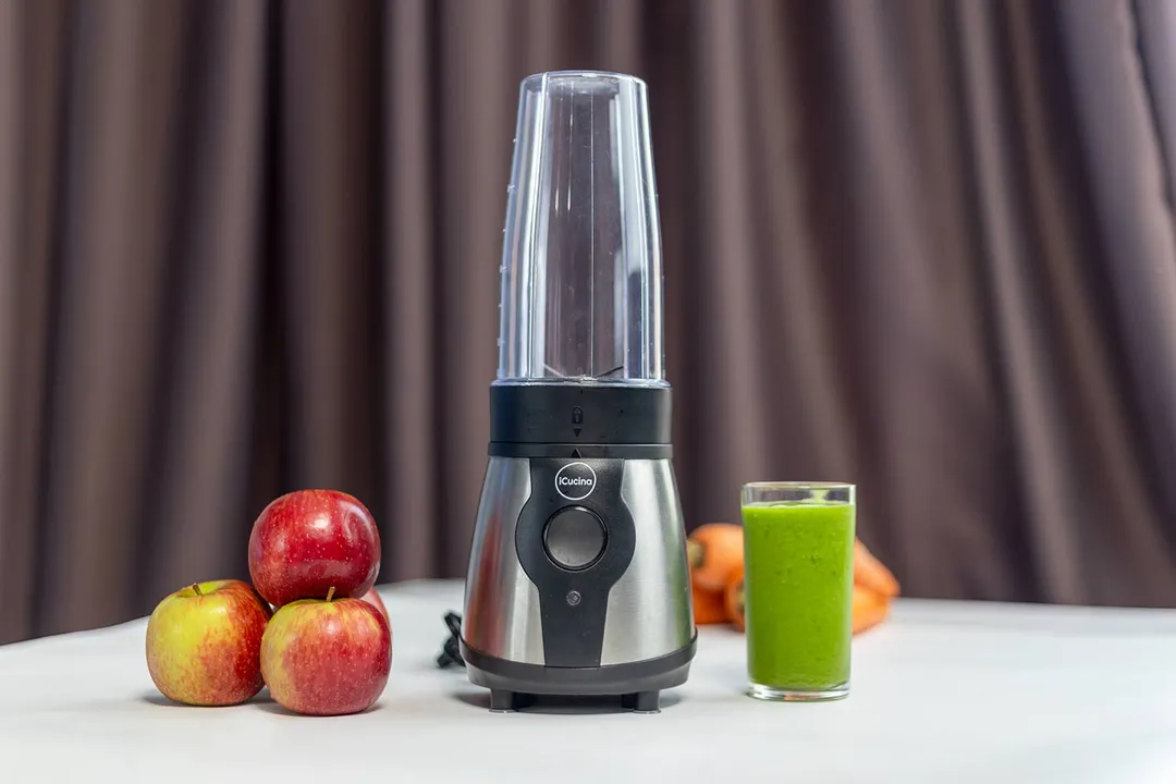 The iCucina personal blender with a cup containing a green smoothie next to some apples and carrots.