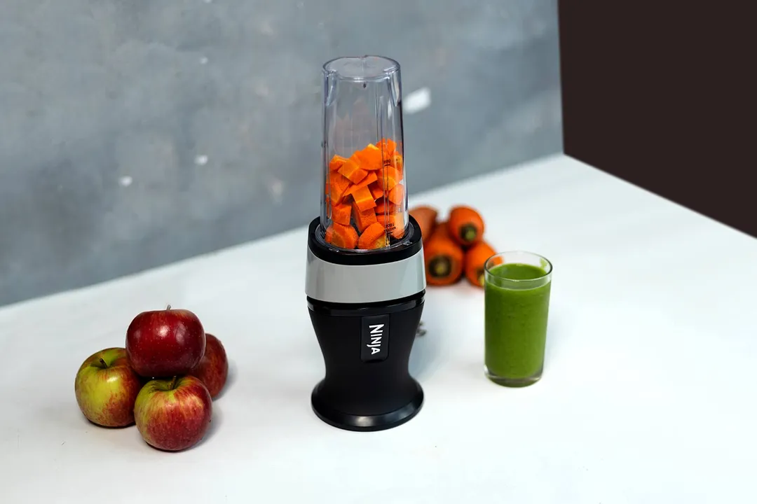The Ninja Fit personal blender with chopped carrots in its cup, next to a glass of green smoothie and whole apples and carrots on a white countertop.