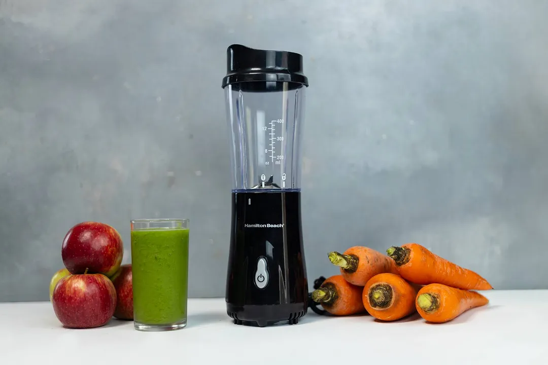 The Hamilton Beach personal blender with a cup containing a green smoothie next to some apples and carrots. 