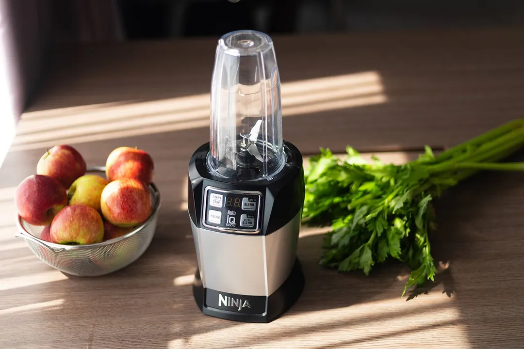 The Ninja BL480D Nutri personal blender on a wooden countertop, next to a bowl of apples and fresh celery.
