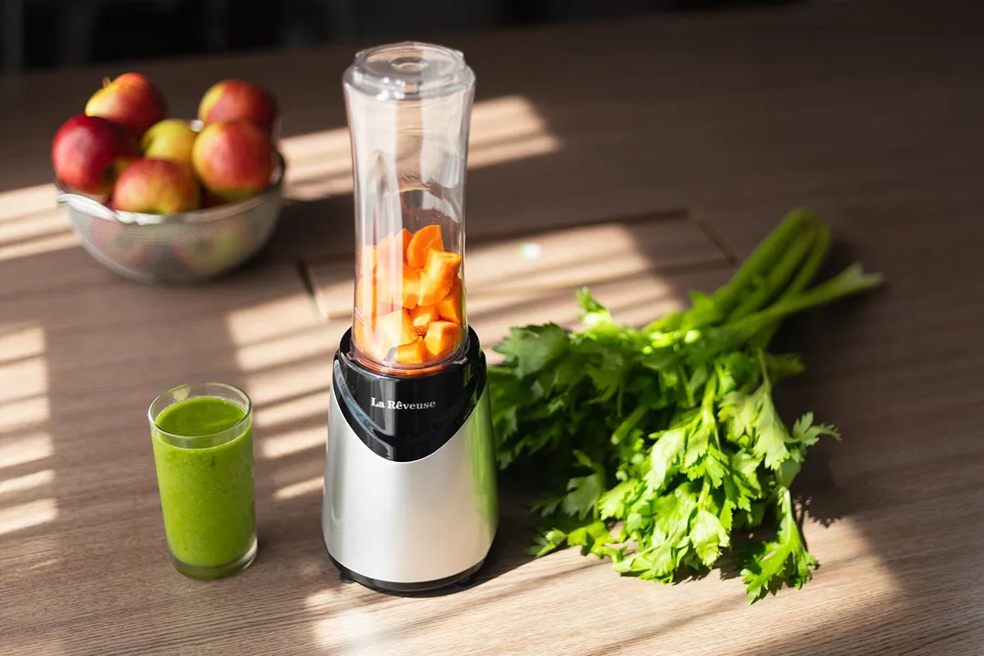 The La Reveuse personal blender with chopped carrots in its cup, alongside a green smoothie and fresh celery, on a sunlit wooden surface.