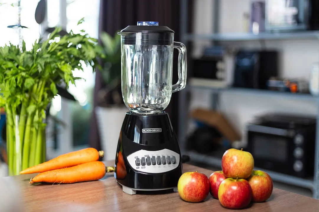 The BLACK+DECKER blender on a kitchen counter with whole apples and carrots nearby, with home appliances in the background.