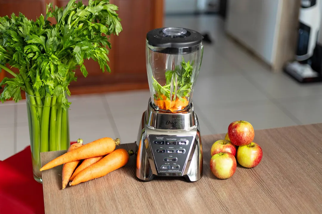 The Oster blender with carrots and parsley prepared for blending, next to apples on a kitchen countertop.