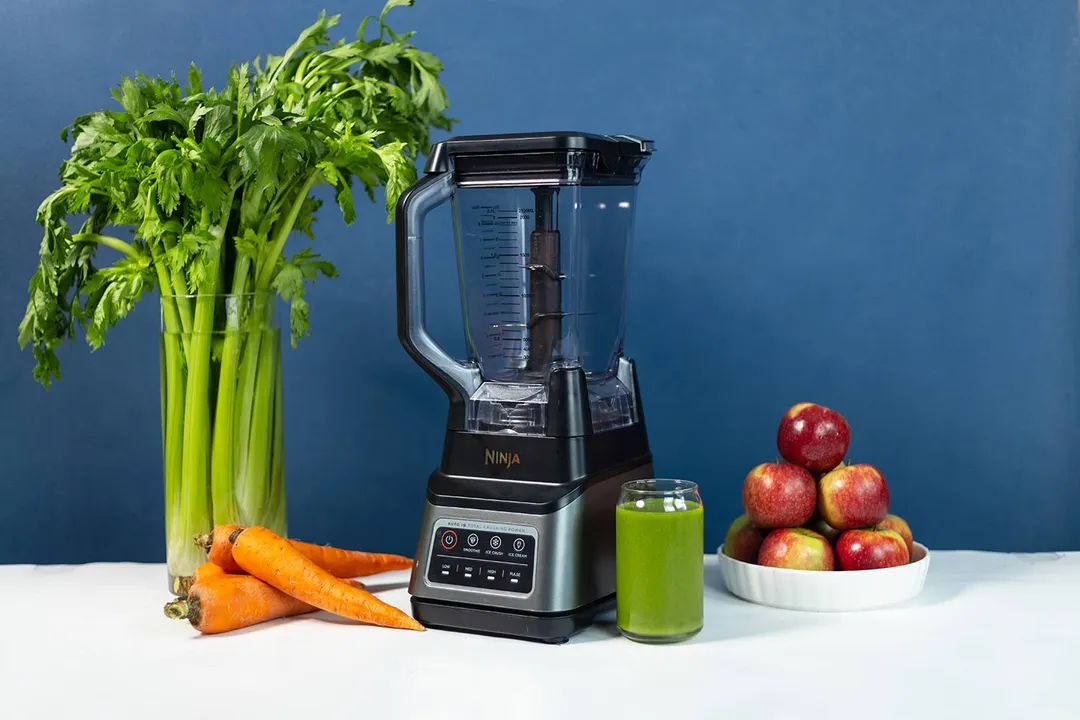 The Ninja BN701 blender on a white surface, next to a bunch of celery, carrots, and a bowl of apples with a green smoothie in front.