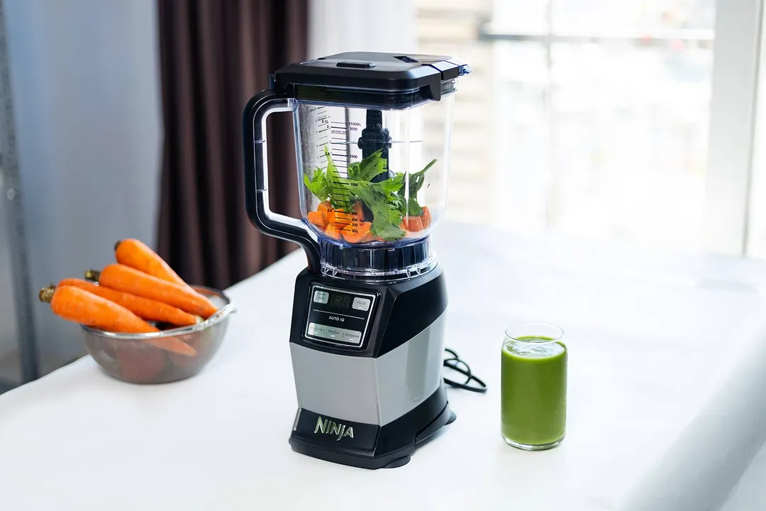 The Ninja AMZ493BRN Compact Kitchen System on a white countertop filled with carrots and parsley, next to a bowl of carrots and a glass of green smoothie.