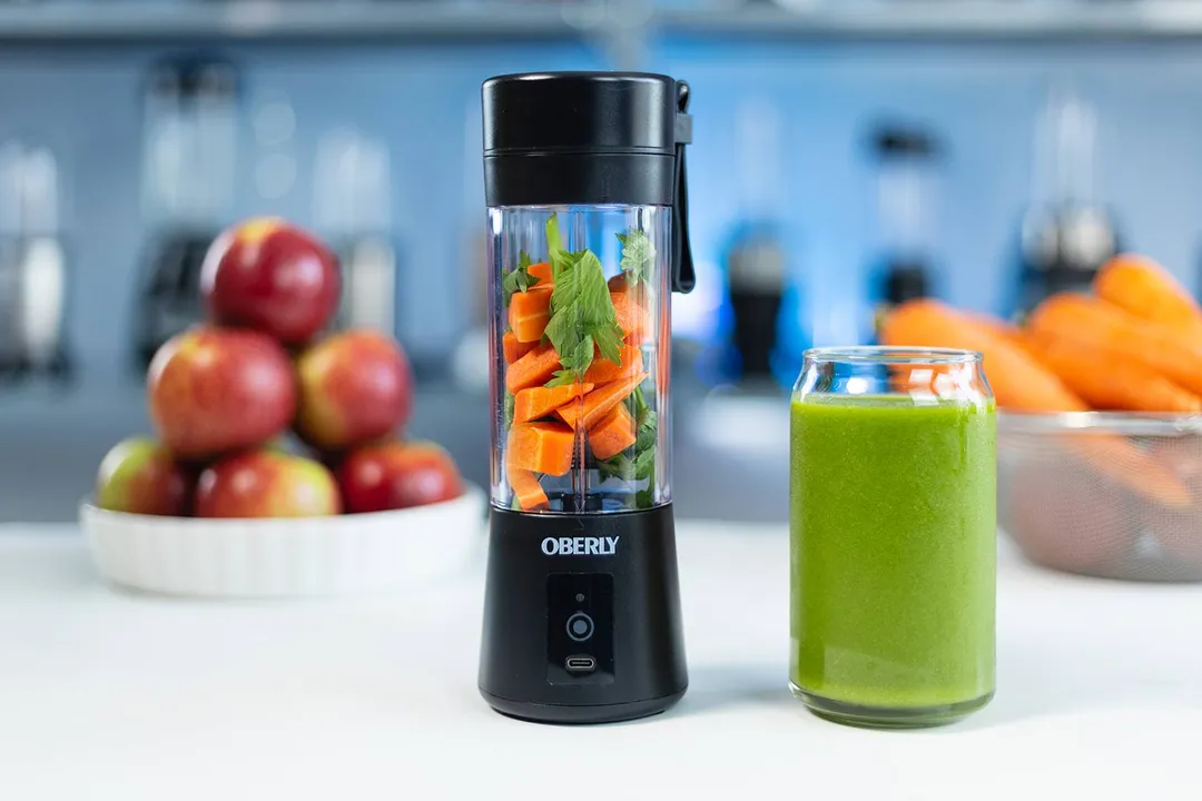 The Oberly cordless portable blender with carrots and parsley inside, placed on a counter next to a green smoothie and a bowl of apples, with kitchen appliances in the background.