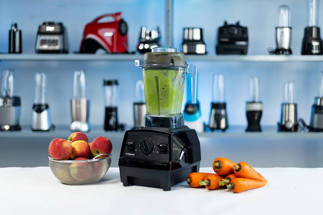 The Vitamix E310 Explorian blender on a countertop with a green smoothie inside, flanked by apples and carrots, against a backdrop of assorted blenders on shelves.