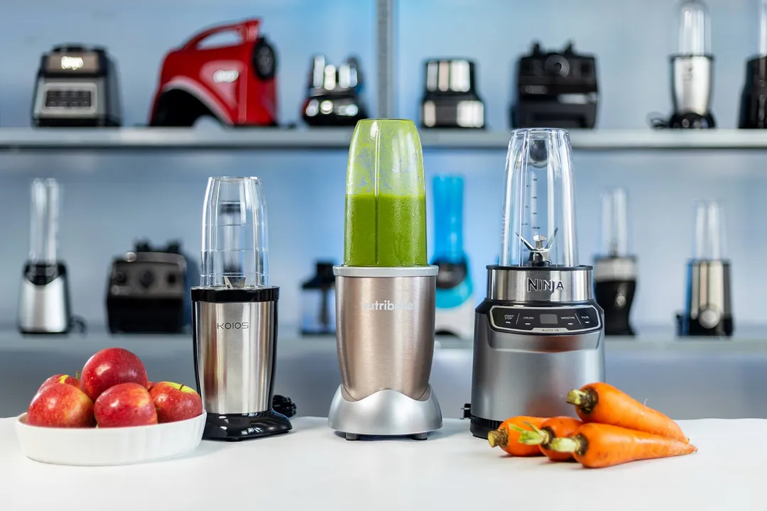 A countertop display of three different personal blenders, including the KOIOS, NutriBullet with a green smoothie inside and the Ninja BN401, flanked by apples and carrots.