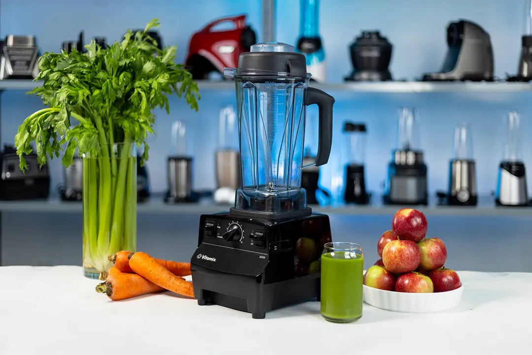 The Vitamix 5200 blender on a countertop with a bunch of celery and carrots on the side, a glass of green smoothie, and a bowl of red apples, set against a blue background with various blenders on display.