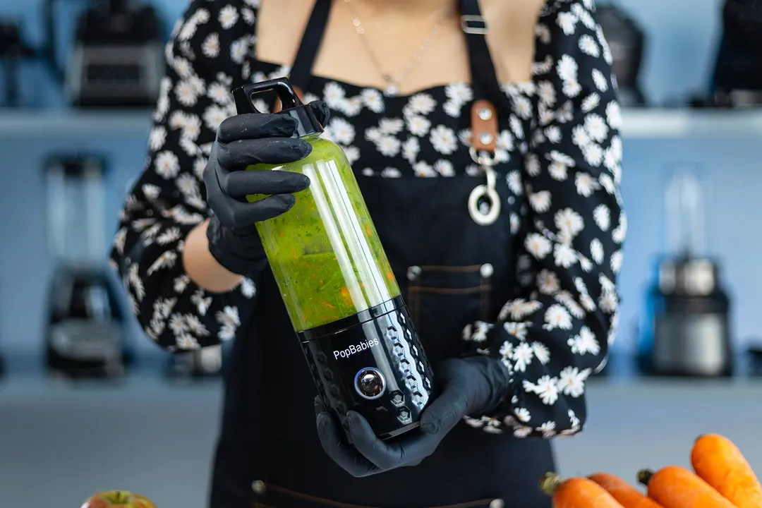 Someone holding the Popbabies cordless portable blender with kitchen appliances in the background.