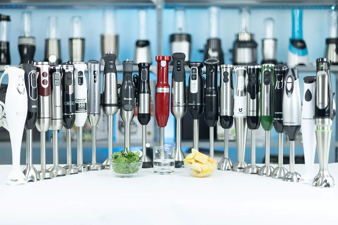 Twenty two immersion blenders standing in a table with the ingredients for the frozen fruit smoothie test, including mango, pineapple, and kale, next to them.  Caption: We put dozens of immersion blenders through the same tests to find the best performing models