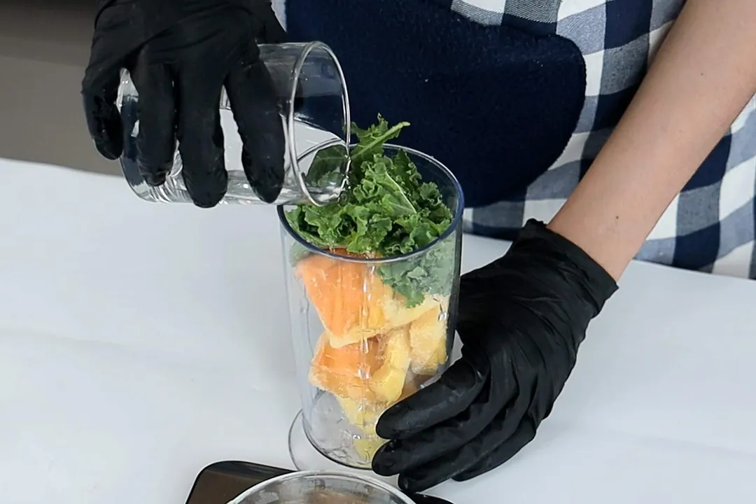 hands pouring water into beaker containing kale, mango, pineapple