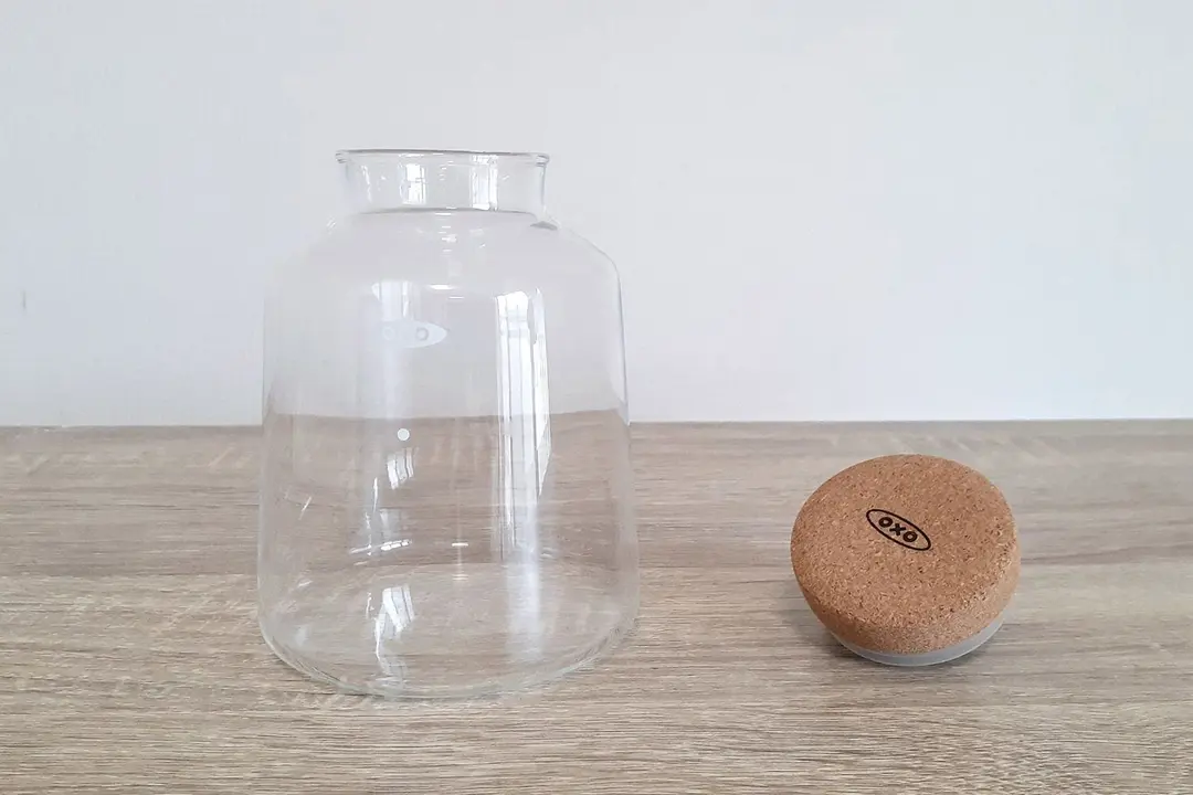 The small glass carafe of the Oxo compact cold brew coffee maker resting on a wood counter with the cork stopper to the right.