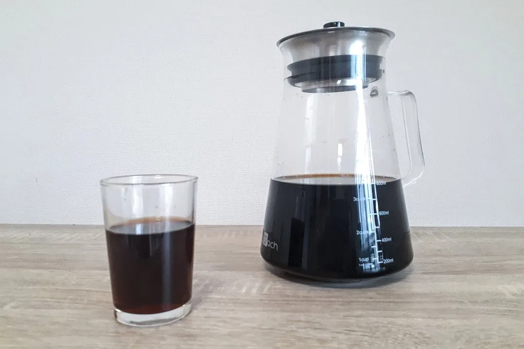 A glass filled with coffee in the foreground and a glass cold brew coffee maker carafe behind it.