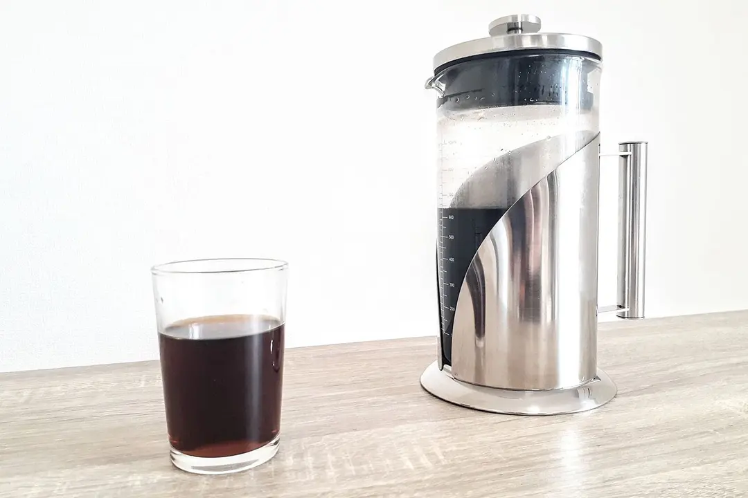 A glass of freshly-poured coffee standing in front of a cold brew coffee carafe.