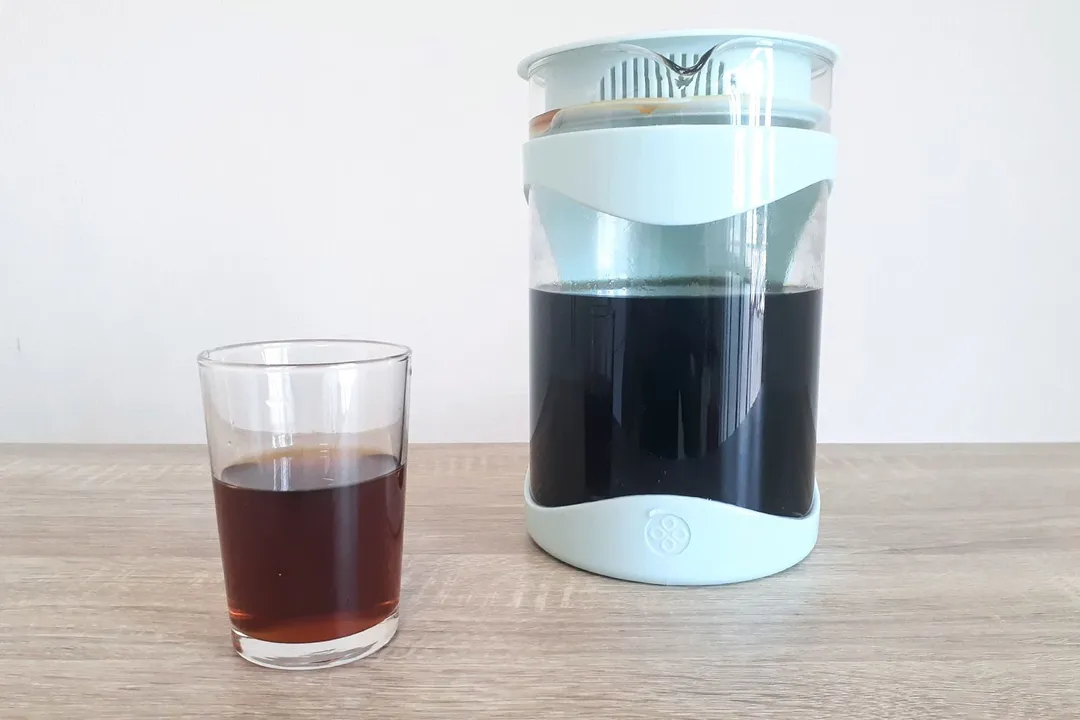 A glass of straight coffee standing in front of a brew carafe of a cold brew coffee maker.