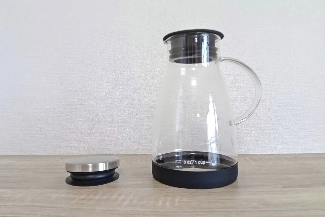 The glass brew carafe of Bean Envy cold brew coffee maker on a wooden countertop with an additional stopper to the side.
