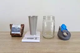 A bag of coffee grounds, a stainless steel filter on a scale, a mason jar, and a lid all ready for making cold brew coffee.