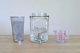 A gauze filter filled with coffee grounds, a 1-gallon cold brew coffee maker half-filled with water, and a measuring jug.