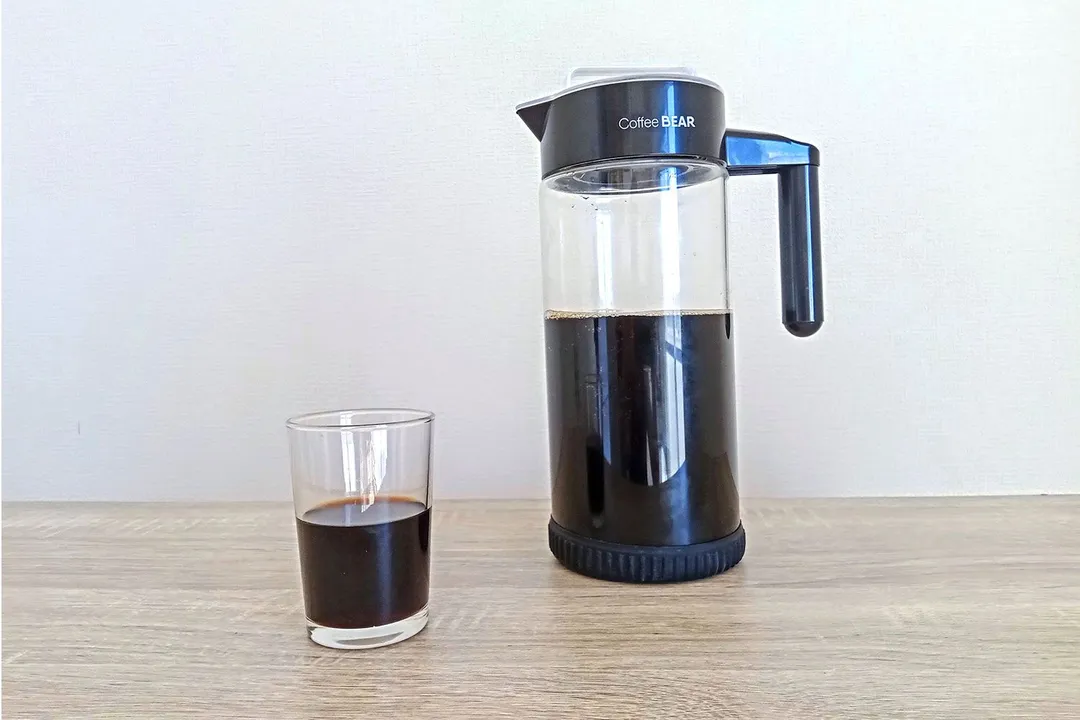 A glass of freshly-poured coffee standing before the Coffee Bear cold brew coffee maker. 