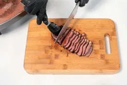On the cutting board, a tong is holding the steak while a knife is slicing the steak.