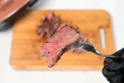 A hand in black glove using a fork to pick up a slice of steak. In the background is the Bella Non-Stick Electric Skillet 14607.