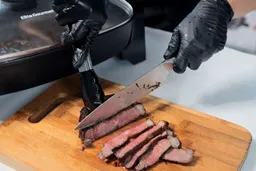 On the cutting board, a tong is holding the steak while a knife is slicing the steak.