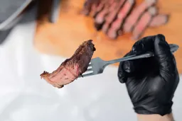 A hand in black glove using a fork to pick up a slice of steak