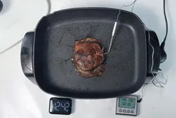 A piece of golden brown steak inside the Elite Gourmet Non-Stick Electric Skillet EG-6203. In front of the skillet is a digital timer and a meat thermometer with its probe inserted into the steak.