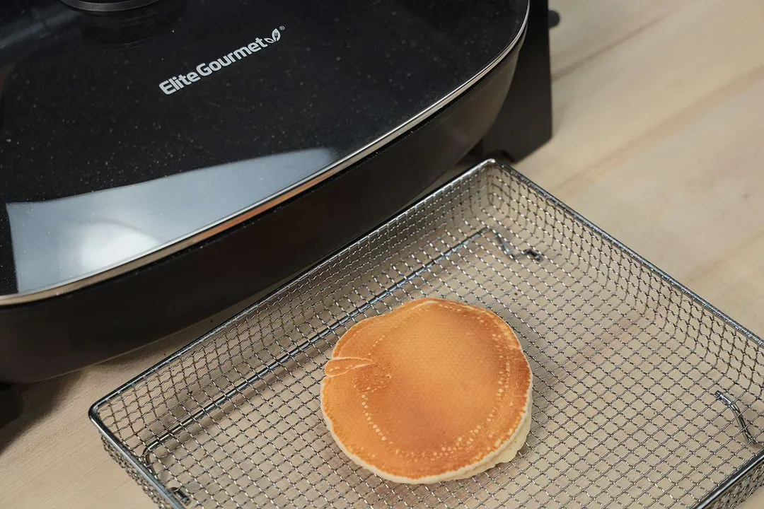 The top side of a golden brown pancake inside an air fryer basket. In the corner is the Elite Gourmet Non-Stick Electric Skillet EG-6203.