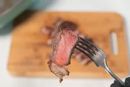 A hand in black glove using a fork to pick up a slice of steak. In the background is the GreenLife Ceramic Non-Stick Electric Skillet CC003725-002.