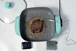 A piece of deep golden brown steak inside the GreenLife Ceramic Non-Stick Electric Skillet CC003725-002. In front of the skillet is a digital timer and a meat thermometer with its probe inserted into the steak.