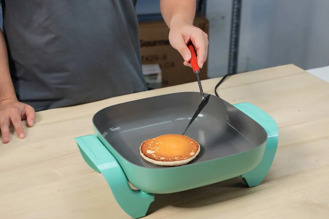 A person holding a spatula flip a golden brown pancake inside the GreenLife Ceramic Non-Stick Electric Skillet CC003725-002.