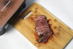 Slices of medium-rare steak on a wooden cutting board. In the background is the Hamilton Beach Ceramic Non-Stick Electric Skillet 38529K.